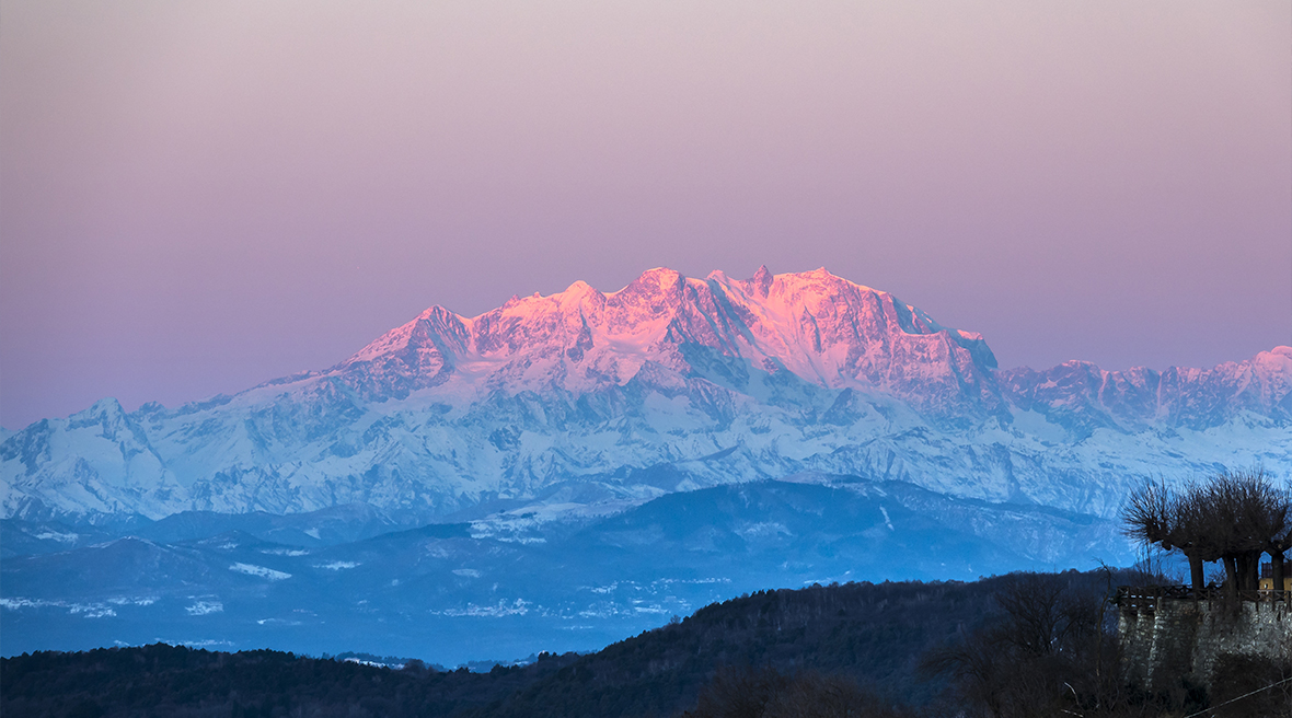 A pink tinted mountain range lit by the sun and rising above trees and valleys below